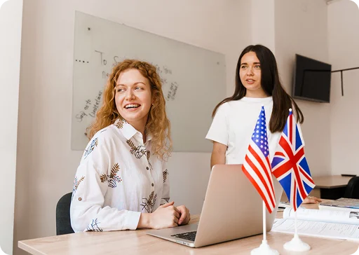 Twee jonge vrouwen in een klaslokaal bij een laptop met de vlaggen van de Verenigde Staten en het Verenigd Koninkrijk op de voorgrond.