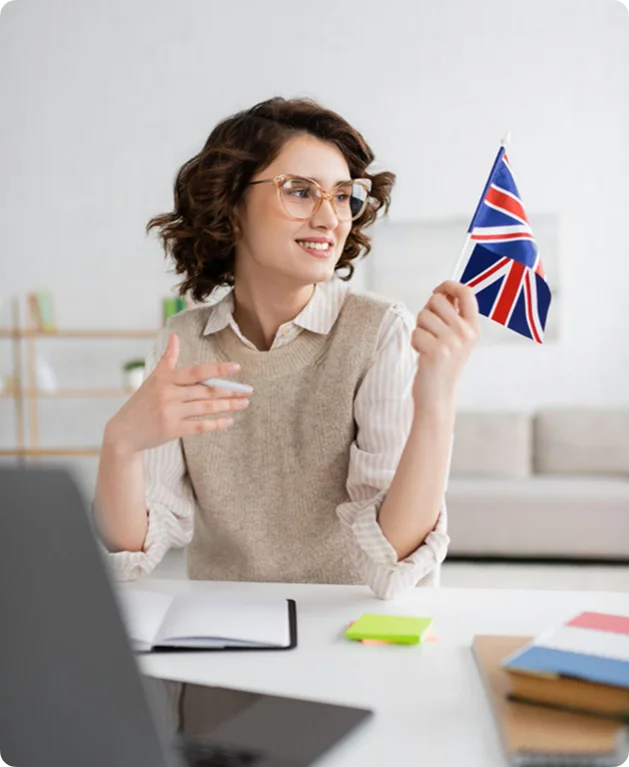 Een jonge lerares met een bril glimlacht en houdt de Britse vlag vast tijdens het geven van een online Engelse les via haar laptop.