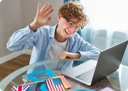 Een vrolijke tienerjongen met een bril zwaait naar zijn laptop tijdens een online les, met kleine vlaggetjes op de tafel.