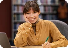 Een glimlachende jonge vrouw zit aan een tafel met een laptop en schrijft in een notitieblok met een bibliotheek op de achtergrond.