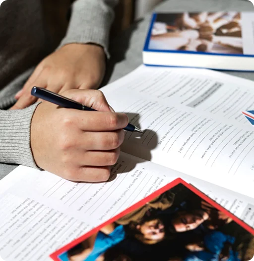 Detailopname van een hand die met een pen oefeningen invult in een Engels werkboek op een tafel.